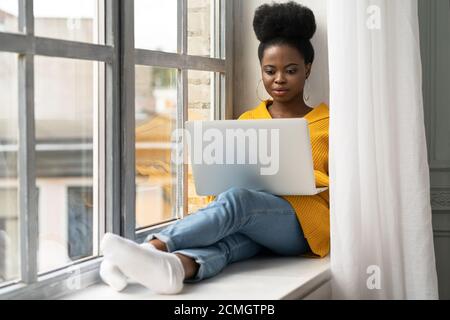 Femme afro-américaine étudiante avec une coiffure afro porter un gilet jaune, assise sur un rebord de fenêtre, travaillant à distance sur un ordinateur portable, apprenant à utiliser onl Banque D'Images