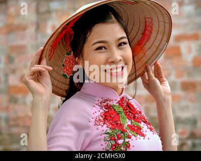 Belle jeune femme vietnamienne tient le bord de son chapeau conique asiatique avec les deux mains et sourires à la caméra. Banque D'Images