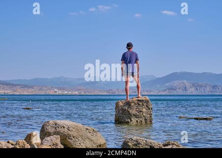 Homme asiatique senior en short, t-shirt et casquette de baseball, tenant dans sa main dans le masque facial, se tient seul sur une pierre dans une baie de mer sur un fond de mont Banque D'Images
