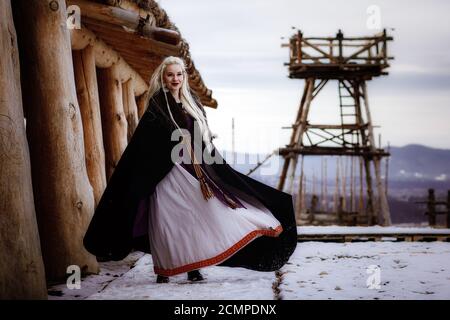 Magnifique viking blond habillé d'un manteau noir sur fond de château. Beau décor de film - un ancien village viking Banque D'Images