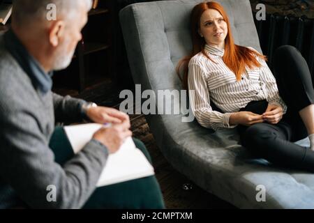 Une jeune patiente heureuse aux cheveux rouges parle de sa santé mentale dans un canapé confortable. Banque D'Images