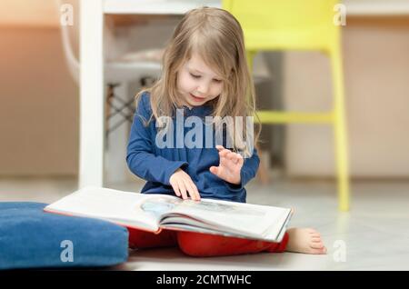 Une petite fille aux cheveux justes examine des illustrations dans un livre pour enfants assis sur le sol à la maison. Développement de la petite enfance Banque D'Images