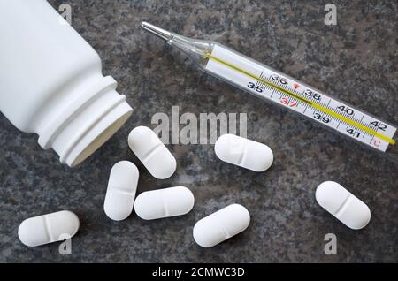 Des pilules blanches et un thermomètre médical au mercure dans le verre indiquant une température critique de 38.2 degrés Celsius sur une surface de table gris foncé. Banque D'Images
