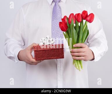 homme dans une chemise blanche et une cravate lilas tenue un bouquet de tulipes rouges avec des feuilles vertes et un cadeau boîte Banque D'Images