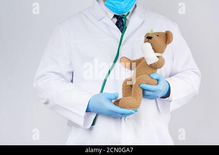 pédiatre en manteau blanc, gants en latex bleu tient un ours en peluche brun avec une patte bannie Banque D'Images