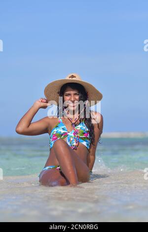 Femme hispanique se trouvant sur la plage à l'intérieur d'une éclaboussure d'eau bleue. Rire naturel tenant chapeau de paille , Beach arrière-plan Los Roques Venezuela Banque D'Images