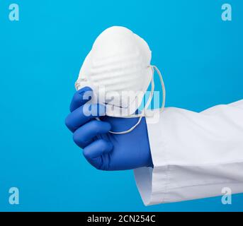 médecin en manteau blanc, gants stériles en latex bleu tient des masques jetables en textile dans la main Banque D'Images