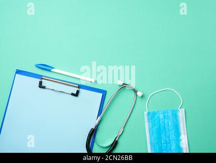 stéthoscope métallique, porte-papier avec une feuille bleue vierge, masque médical bleu jetable sur un bac vert Banque D'Images