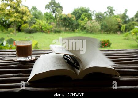 thé dans un verre avec un livre et un spectacle sur une table en bambou dans un beau jardin vert un matin venteux de mousson Banque D'Images