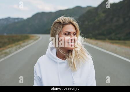 Belle blonde souriante jeune femme voyageur dans blanc à capuche sur la route, voyage à la montagne, Altai Banque D'Images