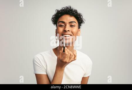 Jeune homme androgyne appliquant du maquillage sur ses lèvres. Gay homme mettant sur le brillant à lèvres avec applicateur contre fond blanc. Banque D'Images