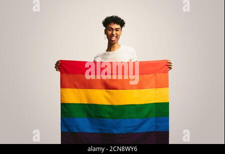 Homme qui dépasse la langue et qui se fait un clin d'œil avec son drapeau de fierté. Homme gay avec le drapeau lgbt faisant un visage drôle sur fond blanc. Banque D'Images