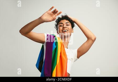 Un jeune homme heureux souriant avec un drapeau de fierté sur fond blanc. Homme fluide de genre avec un drapeau lgbtq sur son épaule. Banque D'Images