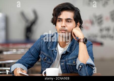 Portrait d'un jeune homme indien beau avec un regard sérieux, perdu en pensée, assis dans une cafétéria de bureau, café, milieu de travail décontracté. Banque D'Images