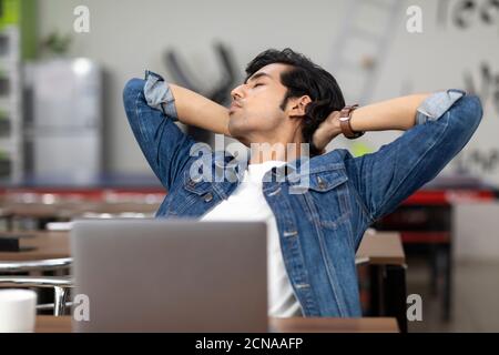 Un Indien fatigué travaillant sur son ordinateur portable, assis dans une cafétéria de bureau, un café, un environnement de travail décontracté, les yeux fermés et se reposant. Banque D'Images