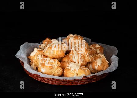 Profiteroles maison à la crème anglaise recouvertes de chocolat blanc fondu dans un panier en osier sur fond noir. Dessert français traditionnel. Touche Bas Banque D'Images
