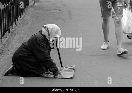 Chernihiv, Ukraine, 7 juin 2019. Une femme âgée dans un foulard supplie pour de l'argent, s'agenouillant dans une rue de ville. Un gars en short passe devant un mendiant. Monochrome Banque D'Images