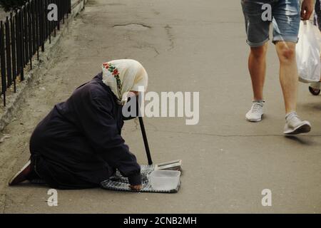 Chernihiv, Ukraine, 7 juin 2019. Une femme âgée dans un foulard supplie pour de l'argent, s'agenouillant dans une rue de ville. Un gars en short passe devant un mendiant. Banque D'Images