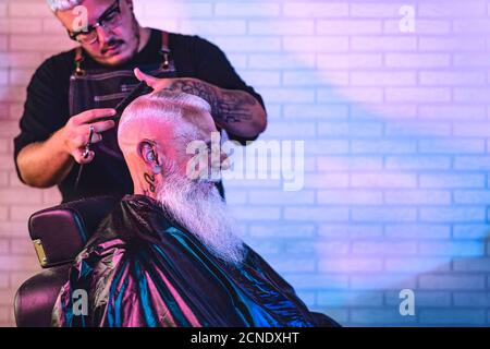 Coiffeur de sexe masculin coupant les cheveux au client principal de l'hipster - côté vue de la jeune coiffeuse travaillant dans un salon de coiffure Banque D'Images