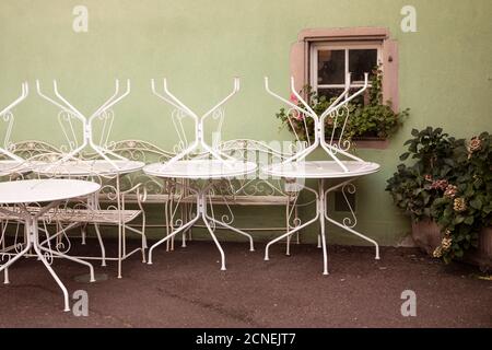 Tables blanches de style vintage à l'envers dans reslauSrant fermé. Mur vert avec fenêtre romantique et plantes vertes. Rue de la vieille ville française. Colmar Banque D'Images