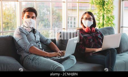 Belle femme asiatique portant un masque d'hygiène médicale protéger faire du travail ensemble dans le salon en utilisant des ordinateurs portables dans la lumière orange de l'heure du soir. W Banque D'Images