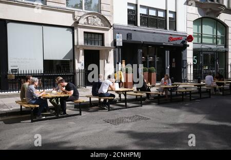 Les gens mangent le déjeuner aux tables et aux bancs placés dans la route adjacente à une baie de stationnement suspendue à Golden Square, Soho, Londres Banque D'Images