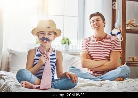 Drôle de temps. Bonne journée pour les pères ! Papa et sa fille jouent à la maison. Jolie fille tient la moustache en papier sur le bâton et fait semblant de papa. Banque D'Images