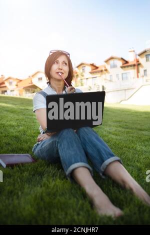 Portrait d'une jolie femme adulte assise sur l'herbe verte dans le parc avec un ordinateur portable sur les jambes, passant la journée d'été travaillant à l'extérieur, en utilisant la Banque D'Images