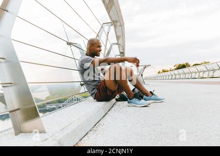 Jeune sportif africain fatigué se détendant après le jogging, tenant une bouteille d'eau sur un pont Banque D'Images