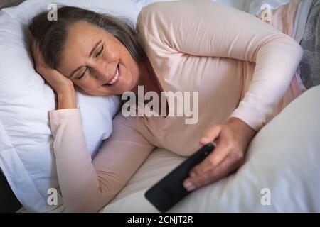 Femme caucasienne appréciant du temps à la maison, distanciation sociale et auto-isolation en quarantaine verrouillage, allongé dans le lit dans la chambre, à l'aide d'un smartphone, smili Banque D'Images