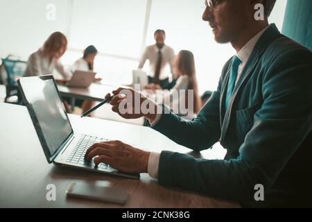 Un homme avec des lunettes travaille dans le bureau derrière un ordinateur portable. Il porte une veste et une cravate. Il y a un stylo dans sa main. En arrière-plan, les collègues du Banque D'Images