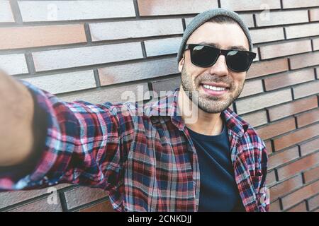 Gai homme élégant en lunettes de soleil et chapeau gris prend un selfie, debout contre le mur de brique marron. Banque D'Images