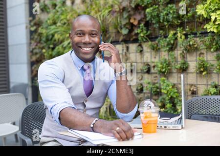 Portrait d'un homme d'affaires noir souriant parlant à travers son smartphone tout en prenant le petit déjeuner. Concept de travail à distance. Banque D'Images