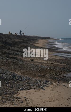 Longue vue sur le rivage en galets avec sable et petites vagues. Chiffres en haut de la banque, quelques pêcheurs. Ciel bleu clair. Format portrait. Banque D'Images