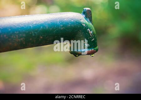 Ancienne pompe manuelle à levier d'eau verte et rouillée avec de l'eau qui coule par le bec verseur, la dernière goutte d'eau, la sécheresse. Arrière-plan vert flou Banque D'Images