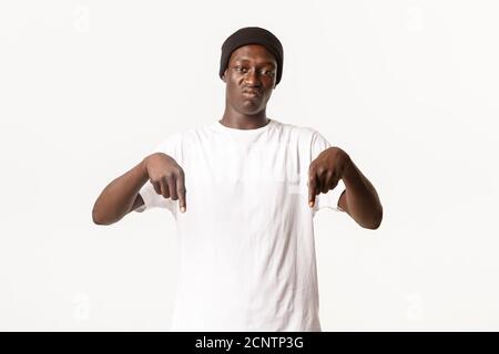 Portrait d'un gars afro-américain sceptique et déçu se plaignant, pointant du doigt vers le bas et grimaçant tracassé, fond blanc Banque D'Images