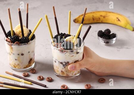 Dessert en verre avec granola, chocolat, banane et mûres décorées de bâtonnets Pocky à la main femelle sur fond gris, gros plan Banque D'Images