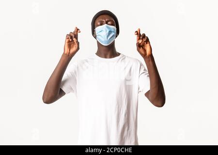 Portrait d'un homme afro-américain nerveux plein d'espoir dans un bonnet et un masque médical, croiser les doigts bonne chance et faire du voeu, debout sur fond blanc Banque D'Images
