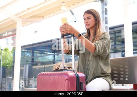 Jeune femme utilisant son smartphone et se reposant sur sa valise, attendant le train à une gare. Concept voyageur. Banque D'Images