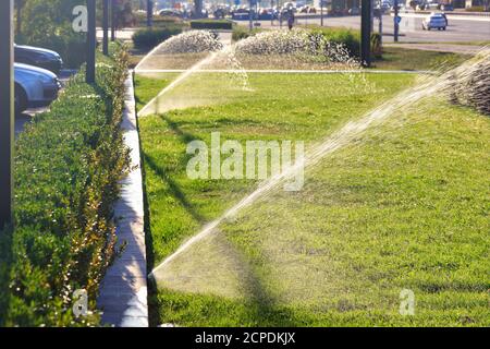 Un système d'irrigation automatique de plusieurs arroseurs irriguaient la pelouse contre un paysage urbain flou par une journée ensoleillée et lumineuse, avec un espace de copie. Banque D'Images