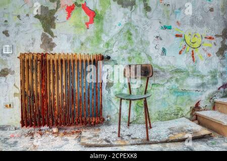 une petite chaise dans un ancien bâtiment abandonné, des gravats et de la poussière, un vieux radiateur rouillé et des dessins d'enfants sur le mur à éplucher Banque D'Images