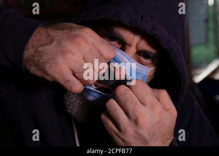 Homme barbu dans un pull à capuche foncé portant un masque chirurgical bleu son visage Banque D'Images