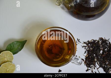 Tasse et théière transparentes avec thé noir, préparation de thé sec sur la table, tranches de bergamote ou citron ou lime sur fond blanc Banque D'Images