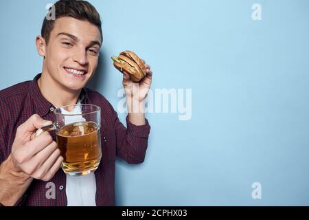 homme gai ivre avec une tasse de bière et un hamburger à la main régime alimentaire style de vie fond bleu Banque D'Images