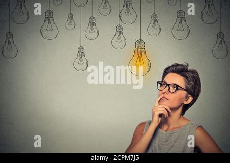 Portrait femme penser dans les verres à la lumière de l'ampoule jusqu'à l'idée au-dessus de la tête isolé sur fond de mur gris Banque D'Images