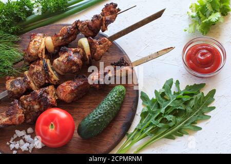 Porc grillé sur brochettes métalliques rôties sur un gril avec des arugula fraîches, du persil, de l'aneth, de la ciboulette sur une planche à découper en bois avec des tomates, du concombre et des tomates Banque D'Images