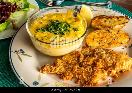 Poisson frit avec sauce, pommes de terre frites, citron et légumes dans une assiette en céramique blanche sur un tapis de table bleu. Banque D'Images