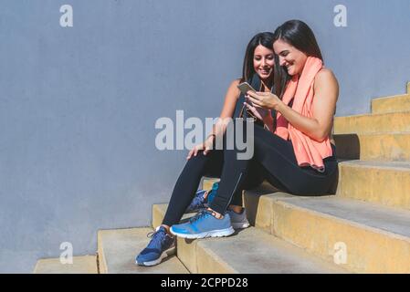 deux jeunes amies aux cheveux foncés portant des vêtements de sport foncés se reposer assis sur les escaliers se montrant les uns les autres téléphone et souriant Banque D'Images