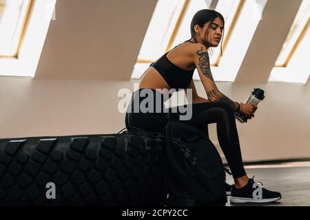 Épuisé sportswoman assis sur un grand pneu dans la salle de gym et contenant une bouteille d'eau Banque D'Images