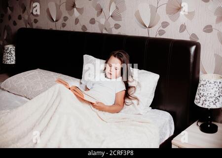 l'enfant est assis sur le lit couvert d'une couverture légère et lit un livre, contes de fées avant d'aller au lit Banque D'Images